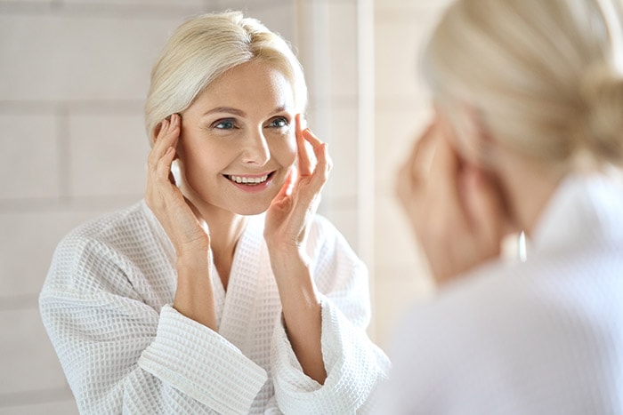 Older woman smiling and touching her face while looking in the mirror, observing Botox effects